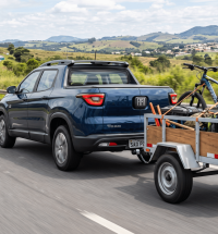 Fiat Toro azul rebocando uma carretinha fazendinha de madeira e metal em uma rodovia, equipada com engate homologado e transportando bicicletas e equipamentos.