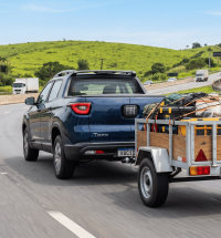 Fotografia lateral traseira de uma picape Toyota Hilux cinza escura rebocando uma carretinha fazendinha de madeira carregada em uma rodovia asfaltada, com colinas verdes ao fundo.