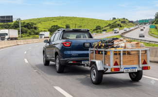Fotografia lateral traseira de uma picape Toyota Hilux cinza escura rebocando uma carretinha fazendinha de madeira carregada em uma rodovia asfaltada, com colinas verdes ao fundo.